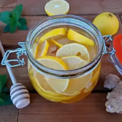 Słoik z wodą, cytryną i imbirem. Obok leżą cytryny, gałązka mięty i drewniana łyżka do miodu.