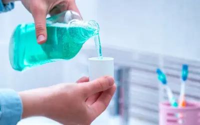 Niebieski płyn do płukania ust jest przelewany z butelki do małego kubeczka. W tle widać szczoteczki do zębów.