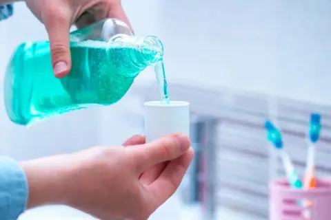 Niebieski płyn do płukania ust jest przelewany z butelki do małego kubeczka. W tle widać szczoteczki do zębów.